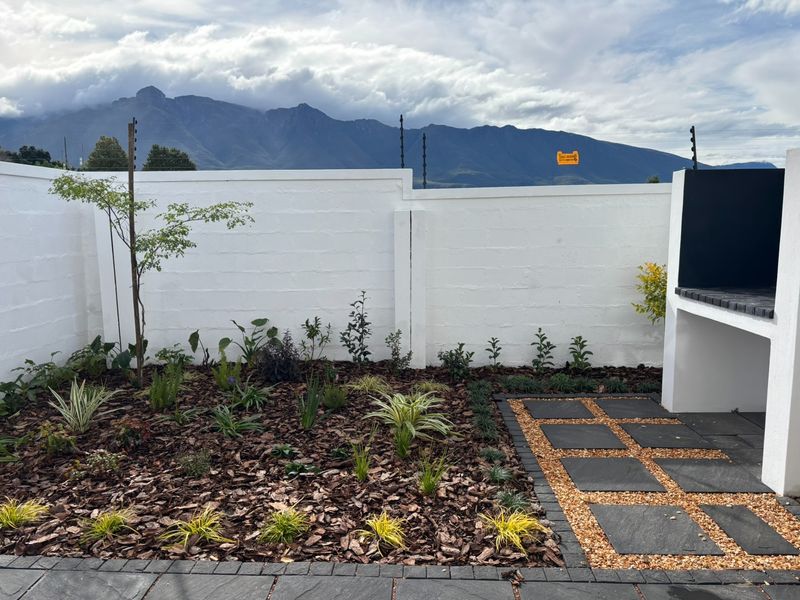 A well-maintained rear garden featuring a paved patio area, built-in braai, and perimeter wall with electric fencing, set against a scenic mountain backdrop.