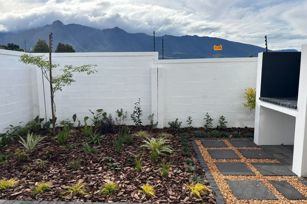 A well-maintained rear garden featuring a paved patio area, built-in braai, and perimeter wall with electric fencing, set against a scenic mountain backdrop.