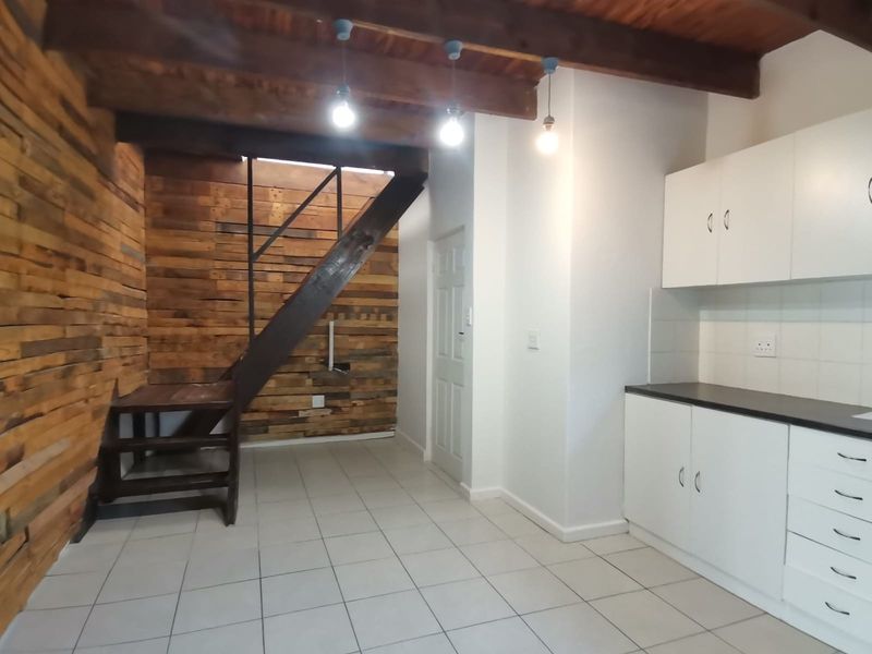 Open plan living area featuring a timber feature wall, exposed ceiling beams, staircase, and a modern kitchen with white cabinetry and tiled flooring.
