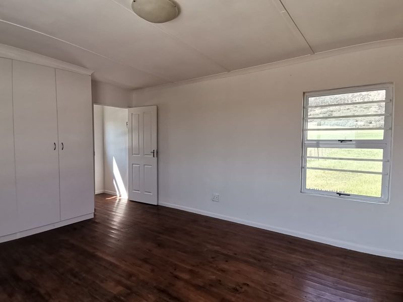 A bright, empty bedroom featuring timber flooring, built-in cupboards, and a louvered window with views of the surrounding landscape.
