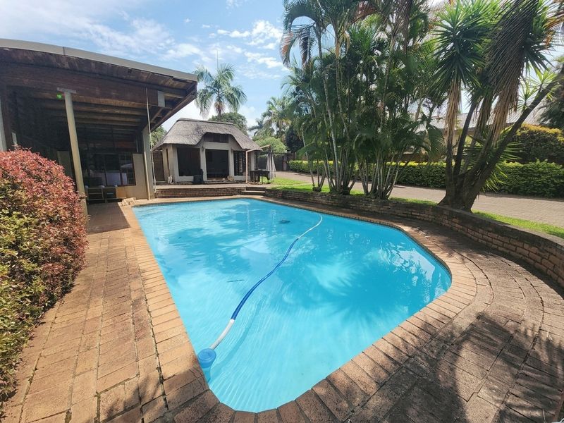 Outdoor pool area featuring a swimming pool, brick paving, and a thatched lapa, set within a landscaped garden environment.