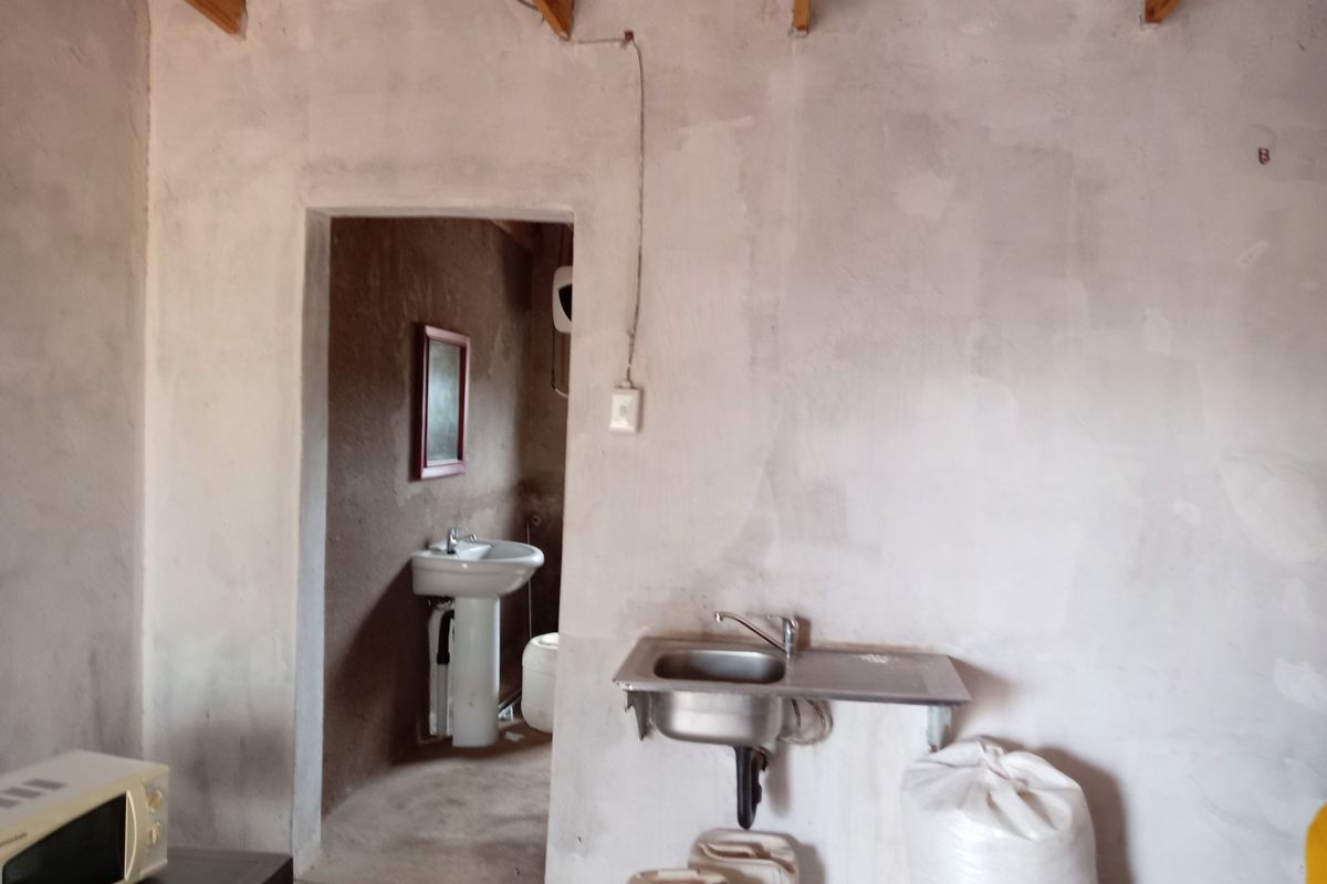 A basic utility or kitchen area featuring an exposed timber ceiling, unfinished walls, a stainless steel sink, and an adjacent room with a pedestal basin.