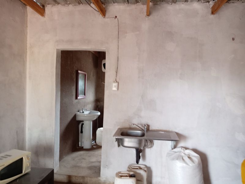 A basic utility or kitchen area featuring an exposed timber ceiling, unfinished walls, a stainless steel sink, and an adjacent room with a pedestal basin.