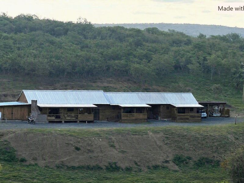 A wide-angle view of a rustic timber-clad farm building featuring a corrugated metal roof, covered verandas, and a brick chimney set in a rural landscape.