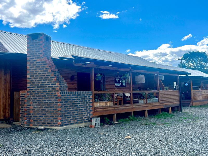 A rustic timber-clad farm dwelling featuring a covered wooden deck and an external brick fireplace, set against a gravel yard.