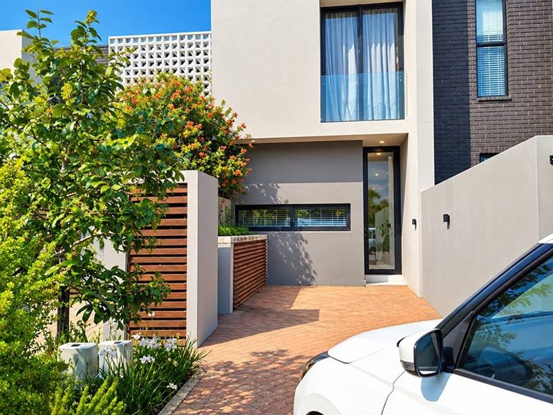 Modern front facade featuring a brick paved driveway, timber slat privacy screening, and contemporary architectural elements including a glass balcony and breeze block detailing.