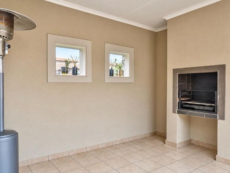 Covered patio area featuring a built-in braai, neutral tiled flooring, and square-framed windows, providing a functional outdoor entertainment space.