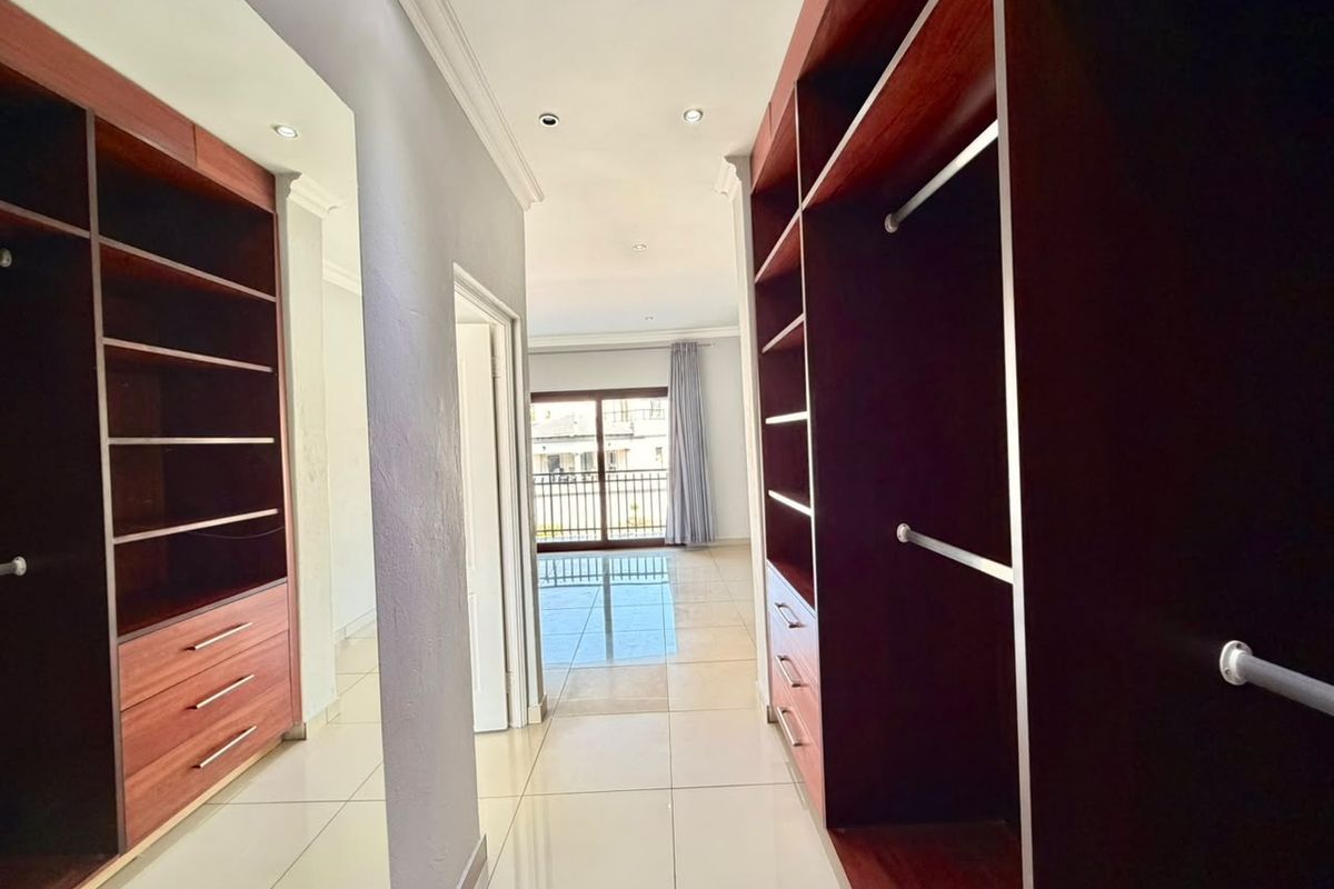A spacious walk-in dressing area featuring extensive built-in wooden cabinetry, neutral tiled flooring, and recessed lighting, leading towards a bright bedroom space.