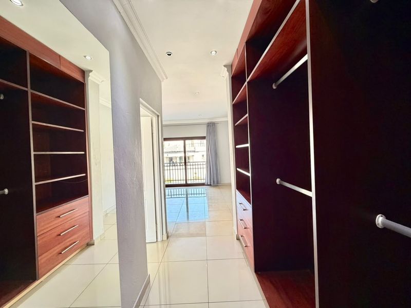 A spacious walk-in dressing area featuring extensive built-in wooden cabinetry, neutral tiled flooring, and recessed lighting, leading towards a bright bedroom space.
