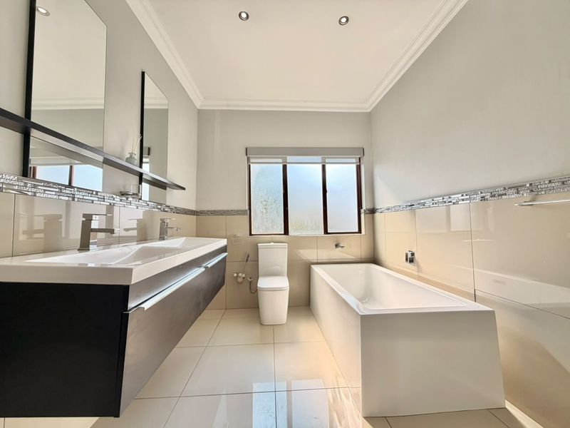 Modern bathroom featuring a double vanity, built-in bathtub, toilet, and neutral tiled finishes with recessed lighting.
