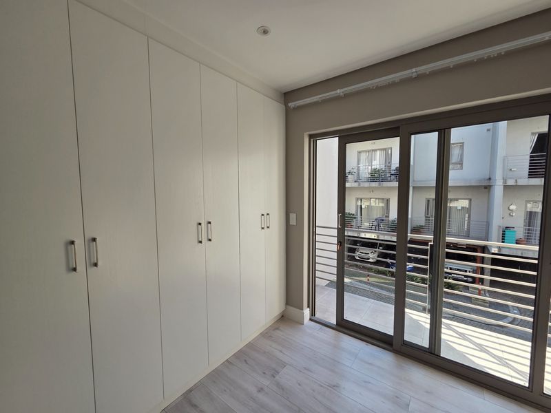 A bright, empty bedroom featuring modern laminate flooring, extensive built-in cupboards, and a sliding glass door providing access to a balcony.