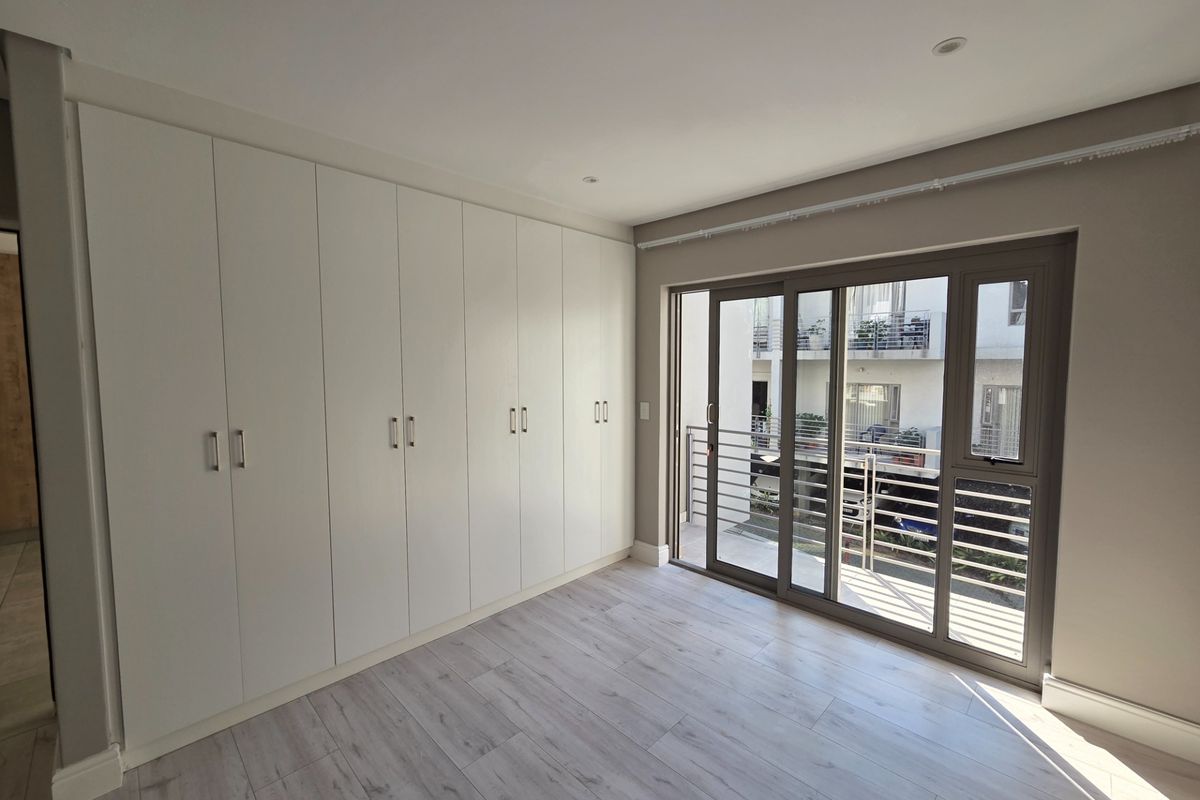 A bright, modern bedroom featuring extensive built-in wardrobes, light-toned laminate flooring, and a sliding glass door providing access to a balcony.