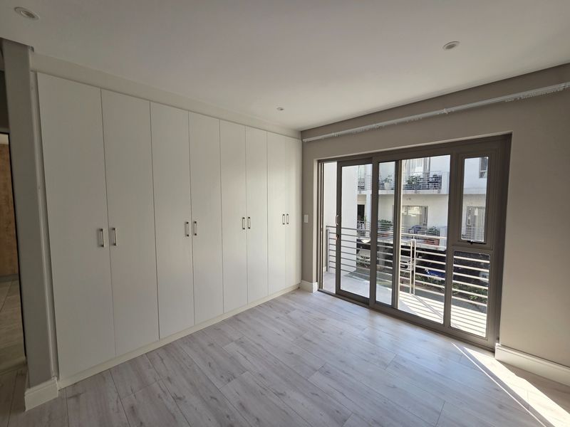 A bright, modern bedroom featuring extensive built-in wardrobes, light-toned laminate flooring, and a sliding glass door providing access to a balcony.