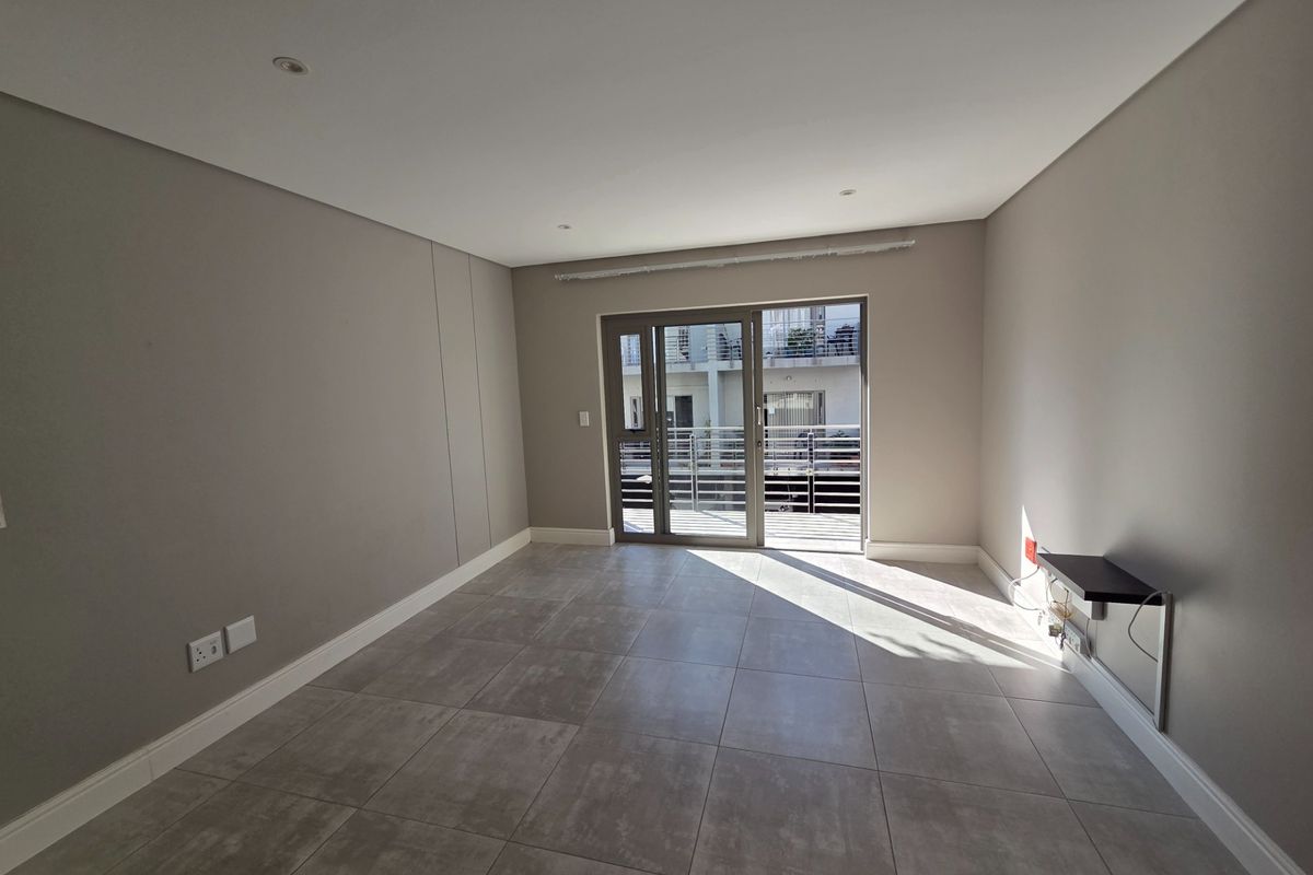 A bright, empty living room featuring modern grey floor tiles, neutral walls, and a sliding glass door providing access to an outdoor balcony area.