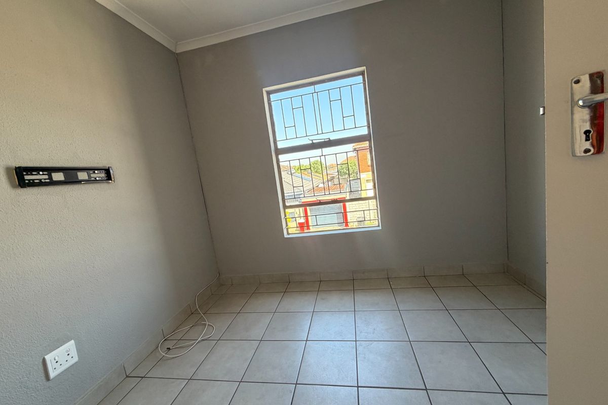 A clean, empty bedroom featuring neutral grey walls, tiled flooring, and a window fitted with security bars.