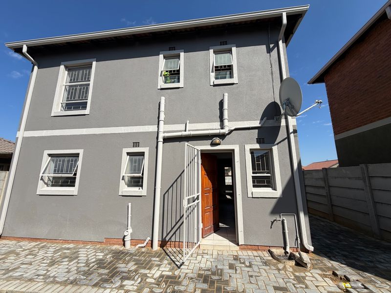 Rear exterior view of a two-story house featuring grey rendered walls, security bars on windows, a paved courtyard, and external plumbing fixtures.