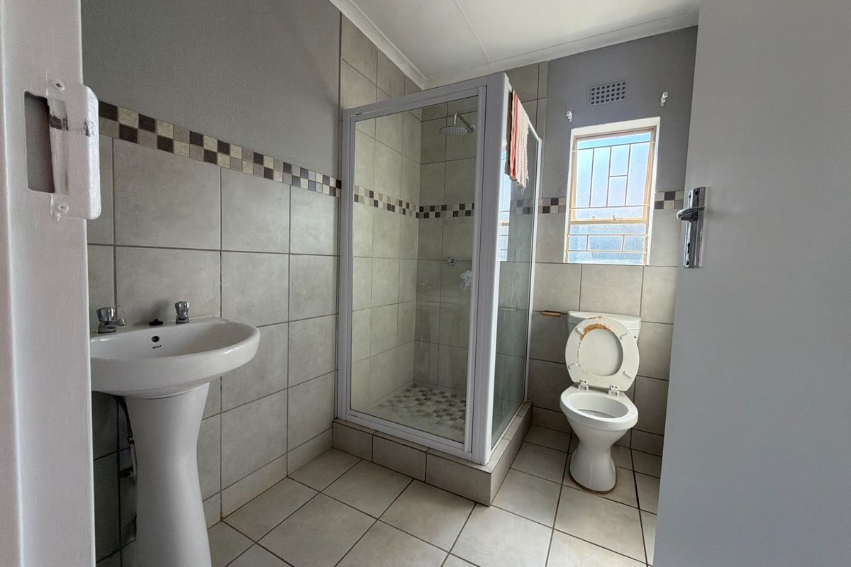 A functional bathroom featuring a pedestal basin, a glass-enclosed shower, and a toilet, finished with neutral wall and floor tiling.