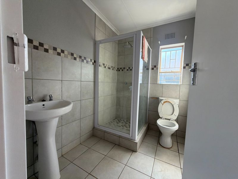 A functional bathroom featuring a pedestal basin, a glass-enclosed shower, and a toilet, finished with neutral wall and floor tiling.