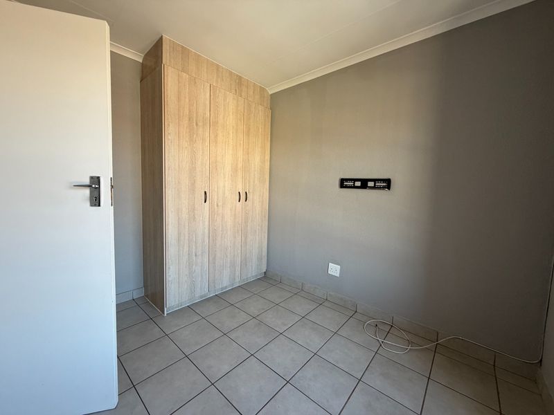 A clean, empty bedroom featuring neutral grey walls, light wood-finish built-in cupboards, and durable tiled flooring.