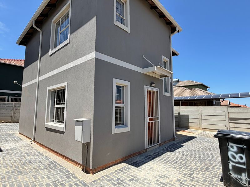 A view of the rear exterior of the two-story house, featuring paved driveway, security gates, and perimeter walling.