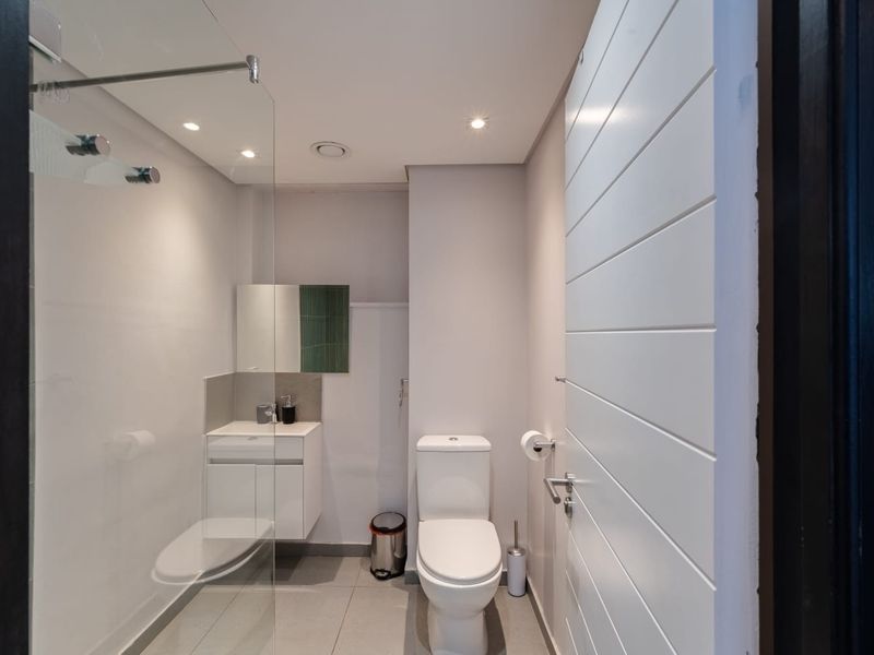 A modern, compact bathroom featuring a glass-enclosed shower, wall-mounted vanity, and contemporary toilet with neutral tiled flooring and recessed lighting.