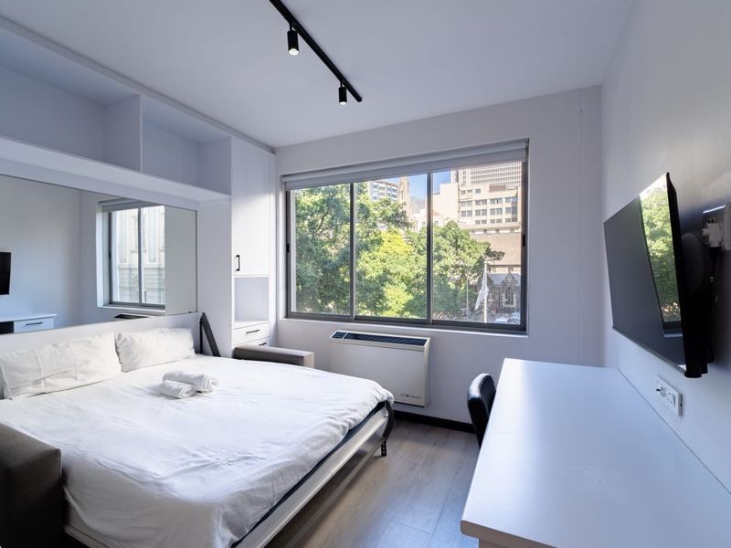 Modern bedroom featuring built-in cabinetry, track lighting, laminate flooring, and a wall-mounted air conditioning unit with large windows offering an urban view.