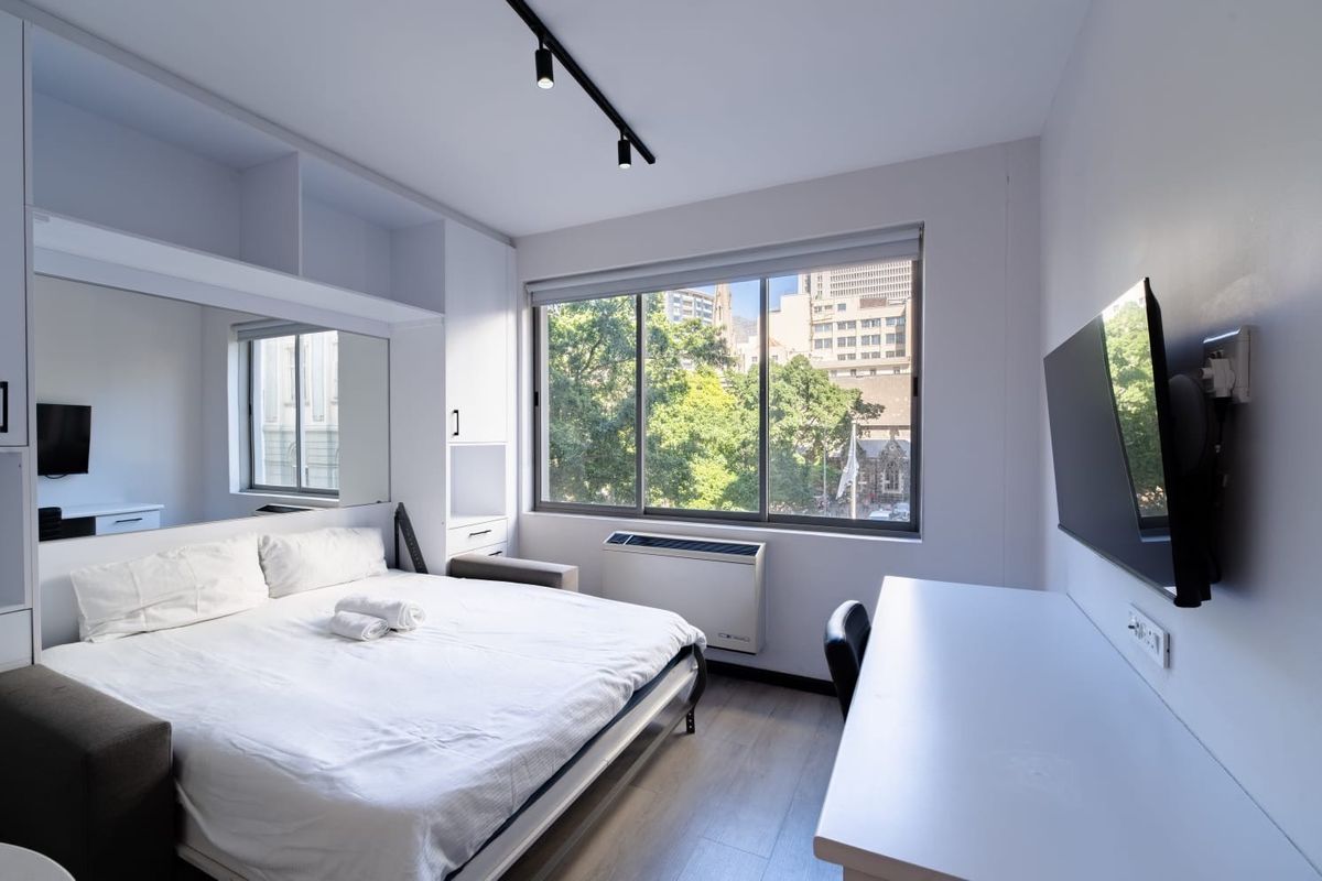Modern bedroom featuring built-in cabinetry, track lighting, laminate flooring, and a wall-mounted air conditioning unit with large windows offering an urban view.