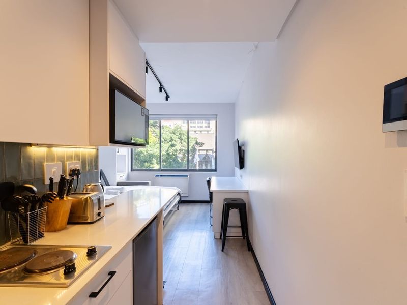 Modern open-plan studio apartment featuring a compact kitchen with built-in cabinetry, electric hob, and a functional integrated desk space with large window views.