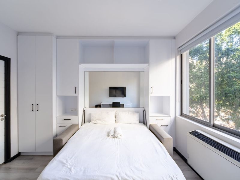A modern, well-lit bedroom featuring extensive built-in white cabinetry, large windows with natural light, and sleek laminate flooring.