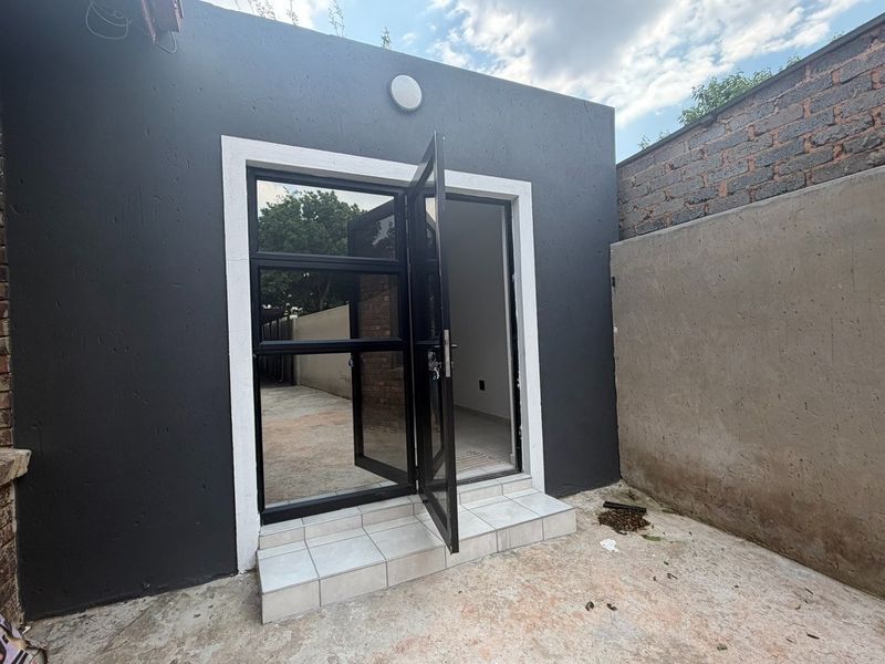 Rear exterior view of the apartment featuring a modern black aluminium glass door, tiled entrance step, and a private concrete courtyard area.
