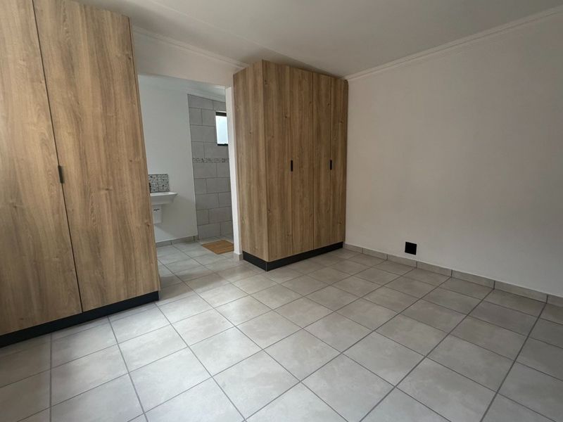 A bright, empty bedroom featuring modern wood-finish built-in cupboards, neutral tiled flooring, and direct access to an en-suite bathroom.