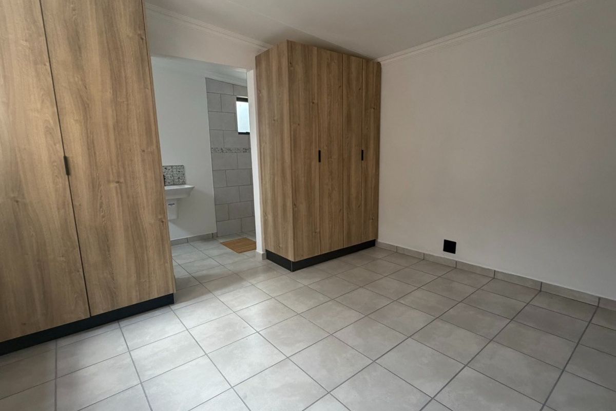 A bright, empty bedroom featuring modern wood-finish built-in cupboards, neutral tiled flooring, and direct access to an en-suite bathroom.