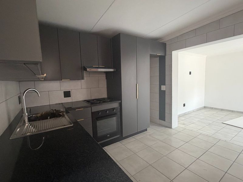 Modern kitchen featuring dark grey cabinetry, stone countertops, built-in oven and hob, and tiled flooring, flowing into an adjacent open-plan living area.