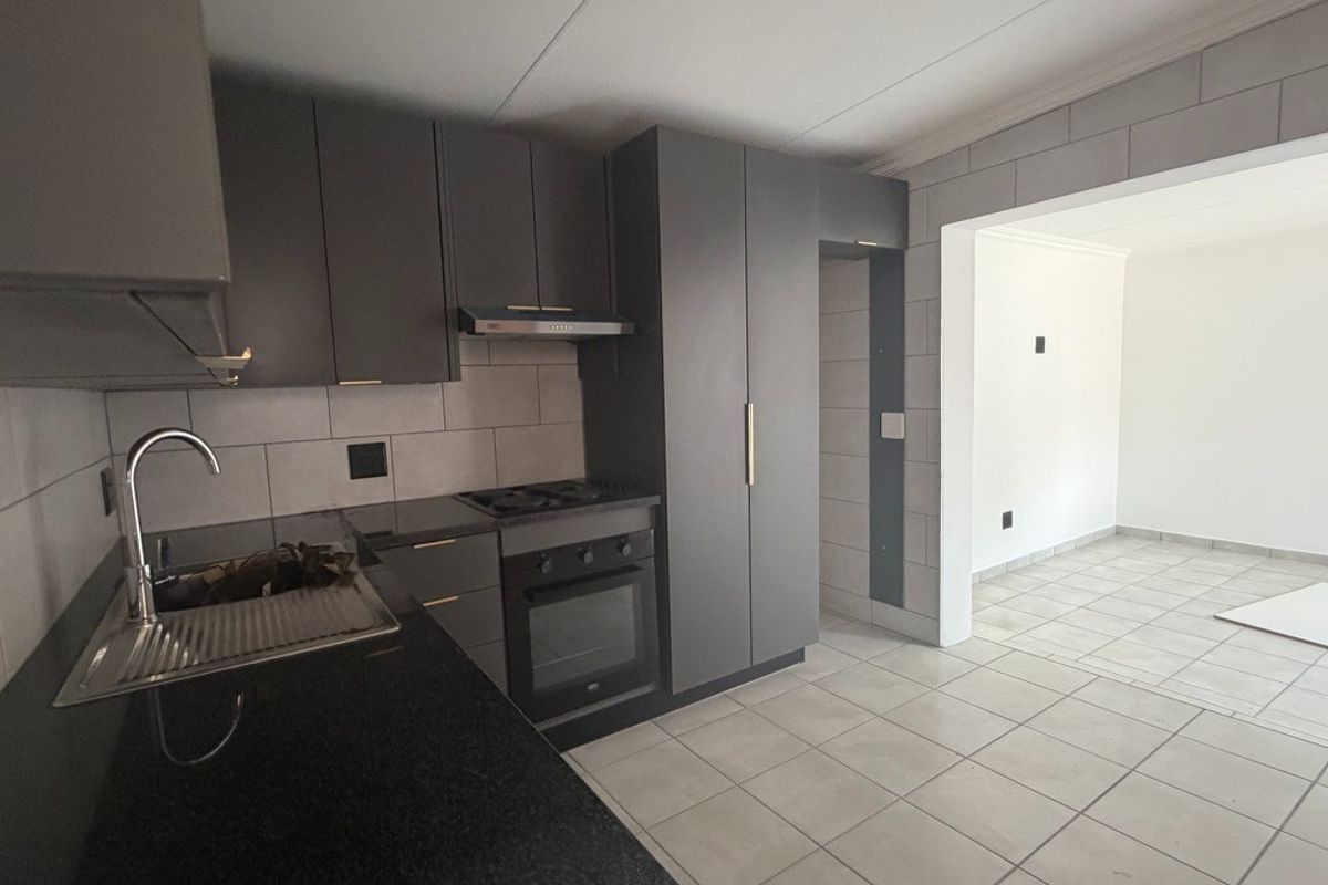 Modern kitchen featuring dark grey cabinetry, stone countertops, built-in oven and hob, and tiled flooring, flowing into an adjacent open-plan living area.