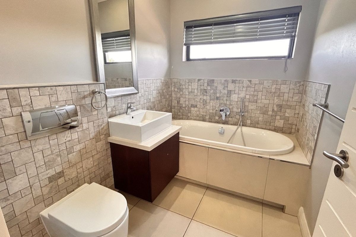 A modern bathroom featuring a built-in bathtub, wall-mounted toilet, vessel sink with vanity cabinet, and neutral stone-look wall tiling.