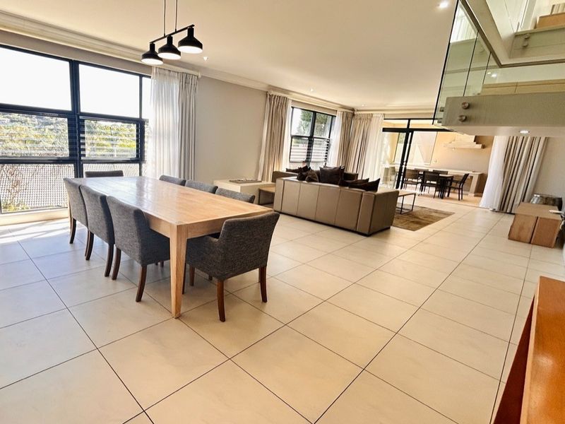 Spacious open-plan living and dining area featuring large windows, tiled flooring, and a modern glass balustrade staircase.