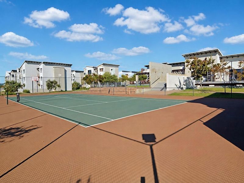 A well-maintained outdoor tennis court with an integrated basketball hoop, surrounded by modern apartment complex architecture and landscaped grounds.