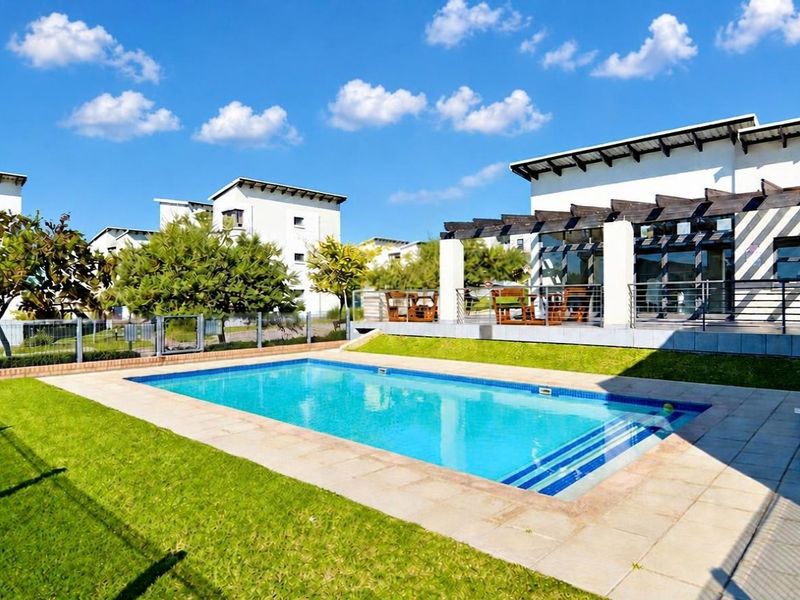 Outdoor pool area featuring a swimming pool with tiled surround, a timber pergola, and a landscaped lawn within a residential complex.