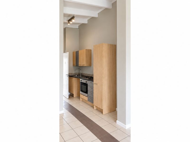 A compact, modern kitchen featuring light wood cabinetry, built-in oven and hob, tiled flooring, and exposed ceiling beams in a clean, neutral space.