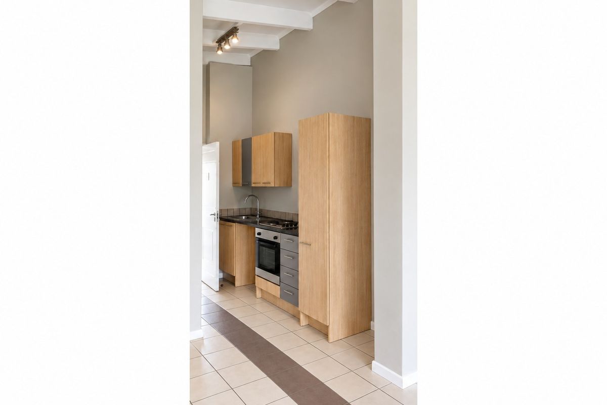 A compact, modern kitchen featuring light wood cabinetry, built-in oven and hob, tiled flooring, and exposed ceiling beams in a clean, neutral space.