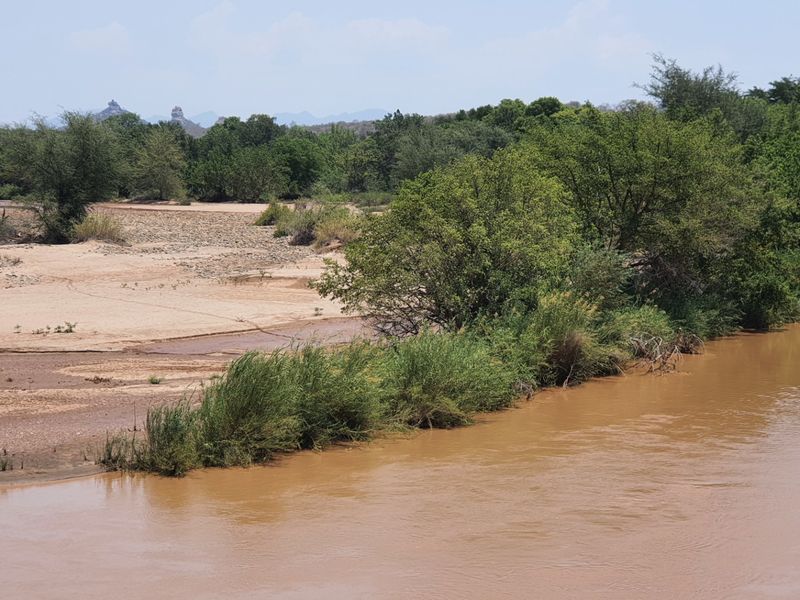 A scenic view of a natural river flowing alongside a sandy bank with lush green vegetation and distant hills.