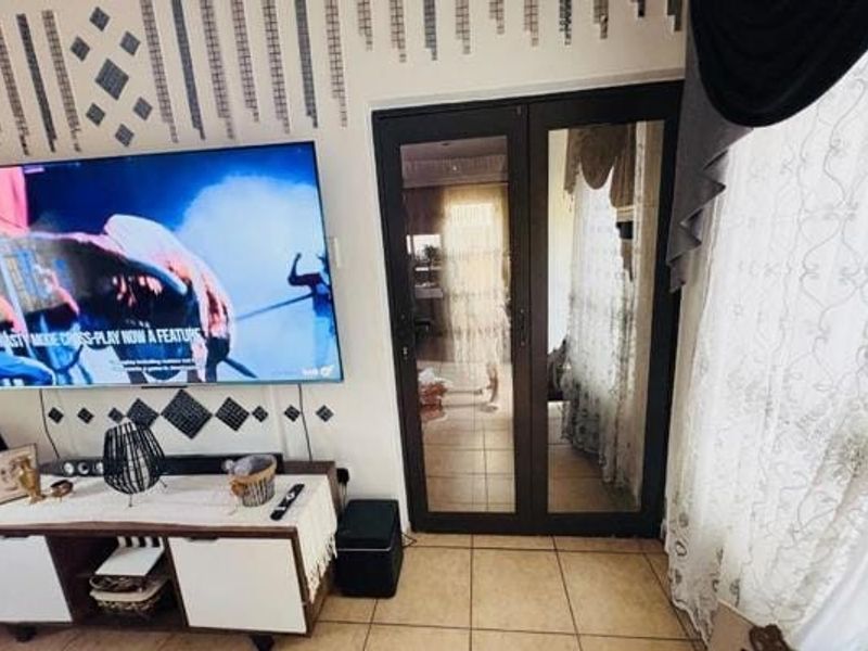 A living room area featuring tiled flooring, a wall-mounted television, and a double glass door leading to an adjacent space.