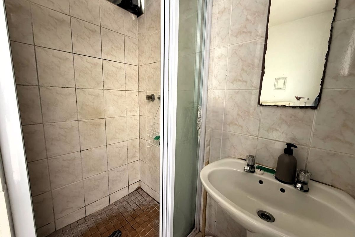A compact bathroom featuring a pedestal basin, a tiled shower enclosure, and wall tiling. The space shows signs of wear and requires maintenance.