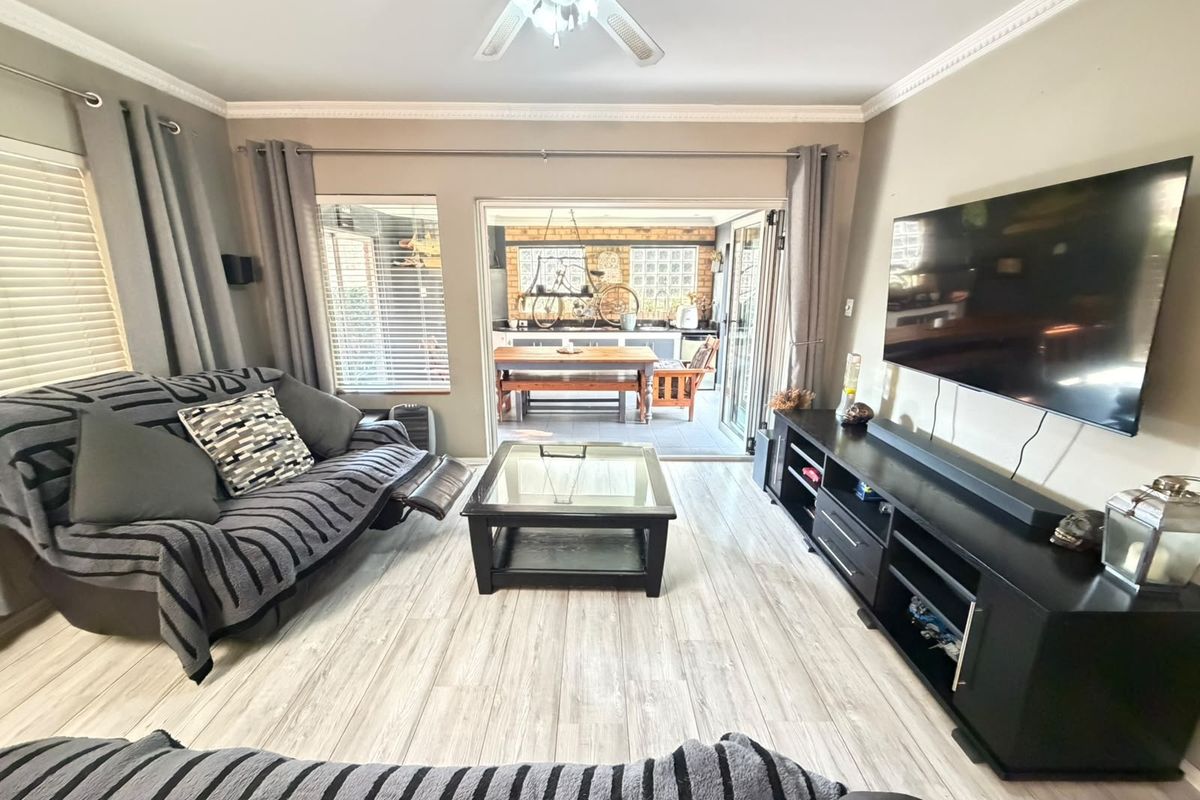 A well-maintained living room featuring light-toned laminate flooring, crown molding, a ceiling fan, and bi-fold doors opening to an outdoor patio area.