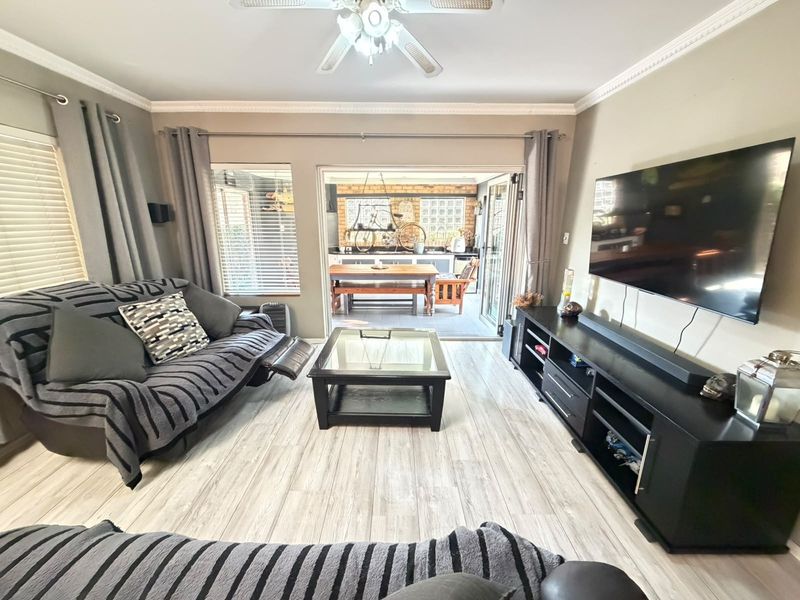 A well-maintained living room featuring light-toned laminate flooring, crown molding, a ceiling fan, and bi-fold doors opening to an outdoor patio area.
