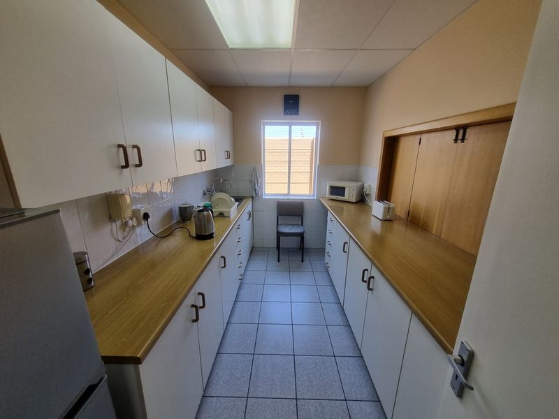 A functional galley-style office kitchen featuring laminate countertops, ample built-in cabinetry, tiled flooring, and a bright window for natural light.