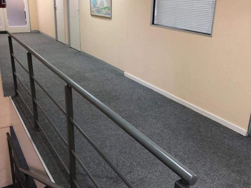 A functional office hallway featuring grey carpeting, a metal safety balustrade overlooking the staircase, and multiple internal office doors.