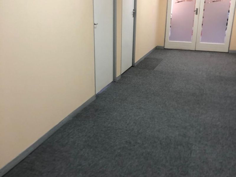 A clean, empty office hallway featuring grey carpet flooring, neutral walls, and glass-panelled double doors leading to an adjacent workspace.
