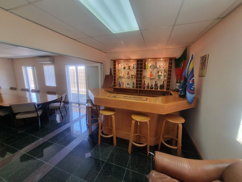 A dedicated office bar area featuring a built-in wooden counter, integrated shelving with display lighting, and polished tiled flooring.
