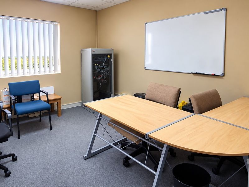 A functional office space featuring a wall-mounted whiteboard, server rack, vertical window blinds, and carpeted flooring.